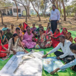 Researchers and female farmers sit on a green tarp during a focus group discussion.